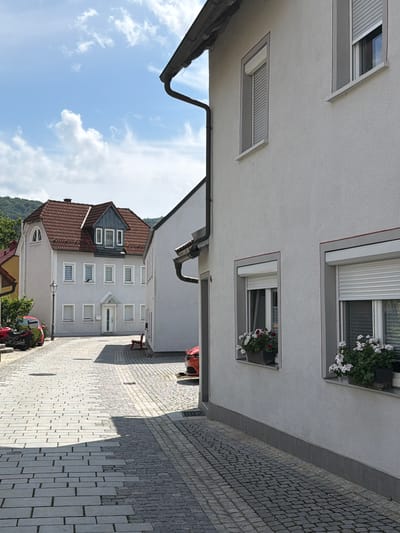 The image shows a street scene in Ebermannstadt, Germany, with a cobblestone road leading towards a building with a red tiled roof. The building on the right is white with a textured facade and has two windows with flower boxes. The street is paved with interlocking cobblestones and has some shadows cast by the buildings. In the background, there is another white building with multiple windows. A red motorcycle and a red car are parked on the left side of the street. The sky is blue with scattered clouds.