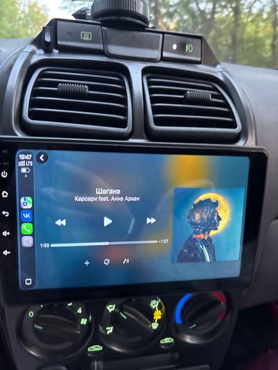In Ladozhskaya, Russia, a car's dashboard is visible, focusing on a large touchscreen infotainment system. The screen displays song information, including the title "Шагане" and artist "Кєрсари feat. Анна Аркан," along with album art featuring a silhouette of a person against a yellow moon. Playback controls, time stamps (1:35 and -1:27), and icons for various apps like VK and a phone are present on the left side of the screen. Above the infotainment system are car air vents and control buttons. Below the screen, manual climate control knobs with indicator lights are visible. The overall environment appears to be the interior of a parked vehicle.