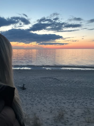 The image shows a beach landscape in Kalngale, Latvia, at sunset. The sky is split between blue and orange hues, with scattered grey clouds. The Baltic Sea reflects the colors of the sunset. The sandy beach is marked with footprints and some driftwood. Blond hair is visible in the foreground. A person is visible at the bottom of the photo wearing a dark shirt.