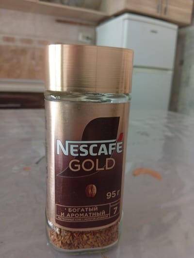 A close-up shot focuses on a jar of Nescafé Gold coffee. The jar, made of glass with a metallic gold lid, is filled with instant coffee granules. The label on the jar is predominantly gold and brown, displaying the Nescafé Gold logo and the text "БОГАТЫЙ И АРОМАТНЫЙ" (Rich and Aromatic) in Russian, along with "95 г" (95g) indicating the net weight. The background shows a blurred kitchen environment with cabinets and a refrigerator. The scene is indoors, on a tabletop, and the lighting appears to be natural, possibly from a window. There are no people visible in the image. The image does not provide any specific details about the city of Chigatay, Uzbekistan, or its features.