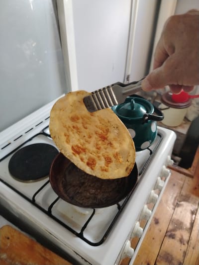 A hand is holding a piece of flatbread with metal tongs over a white gas stove. The flatbread is golden brown and appears to have been recently cooked, with some darker, toasted spots. It is being held above a small, dark pan that is on one of the stove's burners. Next to the pan, another burner is visible. Behind the stove, a green teapot with white polka dots is on the counter, along with two containers of varying sizes, one red and one white. The stove and counter appear to be in a kitchen, possibly in a home. The overall scene suggests food preparation. The location of Shovot, Uzbekistan, is not discernible from the image.