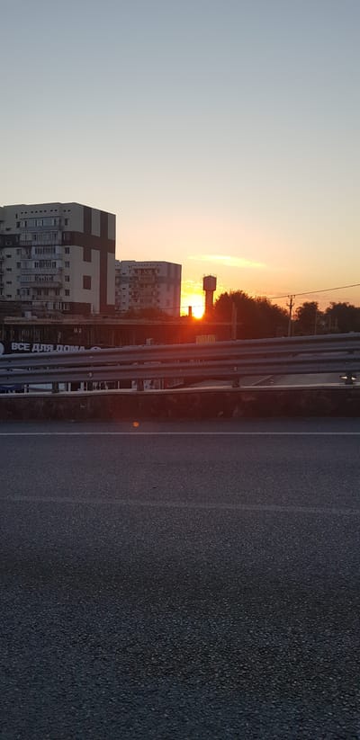The image captures a sunset scene in the Irgeli Rural District, Kazakhstan. The sky is a gradient of pale blue to soft orange and yellow, indicating dusk. In the mid-ground, several buildings are visible. On the left, a tall, multi-story apartment building with a distinctive cross-like dark pattern on its facade stands prominently. To its right, another similar apartment building is situated. A water tower is visible further to the right, silhouetted against the bright sunset. Trees and utility poles are also present in the background. In the foreground, a highway barrier made of metal rails separates the viewer from the buildings. The road surface is dark asphalt with white lane markings. No people are visible in the frame.