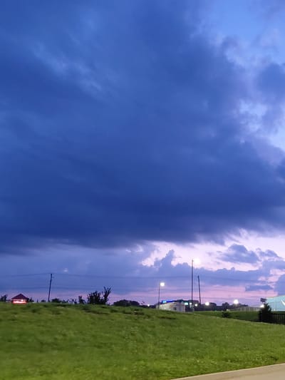 This is a high-angle, medium shot of a grassy hill against a dramatic twilight sky. The sky is a deep, moody blue with large, dark clouds dominating the upper portion of the frame. Lighter, wispy clouds are visible near the horizon, tinged with pink and purple from the setting sun. A line of trees and utility poles runs across the horizon. In the distance, on the left side of the frame, a small building with a red roof and lit signage is visible. On the right side, there are lights from buildings and streetlamps. The grass in the foreground is a vibrant green. The overall impression is one of a storm approaching or dissipating over a suburban or semi-rural area. The location is identified as Lexington, United States of America.