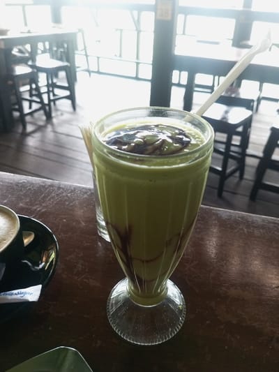 A tall glass of light green smoothie sits on a dark wooden table. The smoothie has a swirl of dark brown chocolate on top and streaks down the side. A white straw and a few unidentifiable objects are in the background. A black cup and saucer are on the left side of the image. The background shows a bright, light-filled outdoor space with tables and chairs.