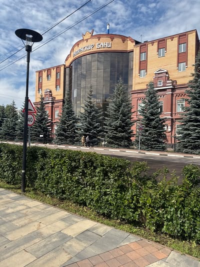 The image depicts a street scene in Ramenskoye, Russia, on a sunny day. A modern building with a glass facade and yellow brick accents stands prominently on the left side of the frame. The building features the text "РАМЕНСКОЕ БАНК" (Ramenskoye Bank) in large, white lettering above the glass section. Several tall, dark green coniferous trees line the front of the building, creating a natural buffer between it and the paved sidewalk.

A paved road runs alongside the sidewalk and the trees. A speed limit sign indicating "40" is visible on a pole near the road. In the distance, along the edge of the road, two people can be seen walking. Further down the street, a lamppost with a dark circular shade is positioned in front of a cloudy blue sky. The sidewalk is paved with interlocking concrete stones in shades of gray and reddish-brown. Lush green bushes and some patchy grass grow between the sidewalk and the road, as well as along the edge of the building. The overall impression is one of a well-maintained urban environment.