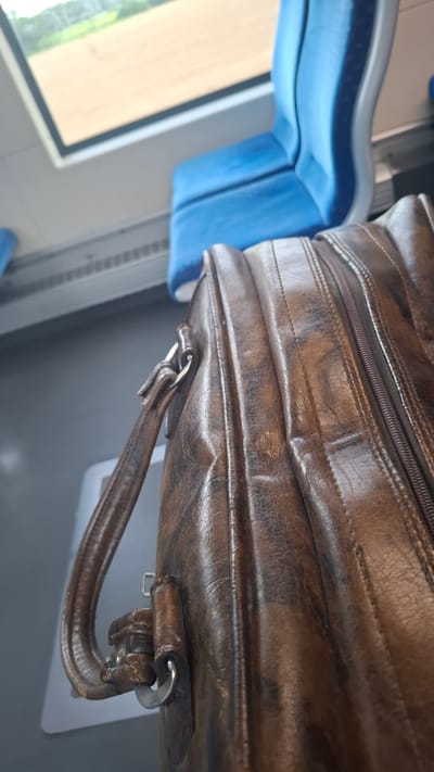 A brown leather briefcase rests on the floor of a train car, positioned in front of blue upholstered seats. Through the train window, a blur of brown and green fields and trees is visible. The scene is indoors, within a public transportation vehicle. The location is identified as Quickborn, Germany.