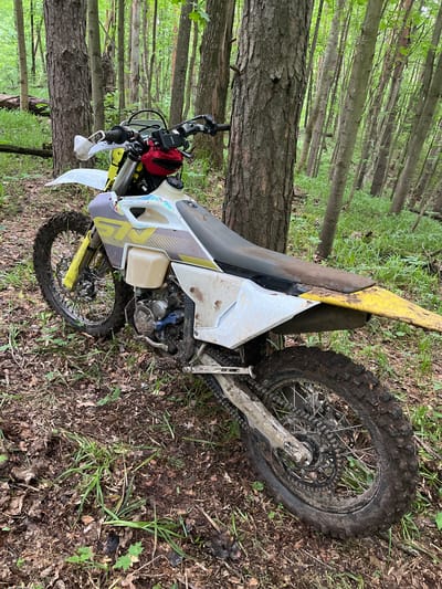 The image shows a dirt bike lying on its side in a wooded area. The bike is white and yellow, and appears to be a Husqvarna. The front wheel is turned slightly towards the left. The tires are muddy. A red pouch is attached to the handlebars. The background consists of trees and undergrowth. The location is not identifiable as Gruzinskii, Russia, from the image.
