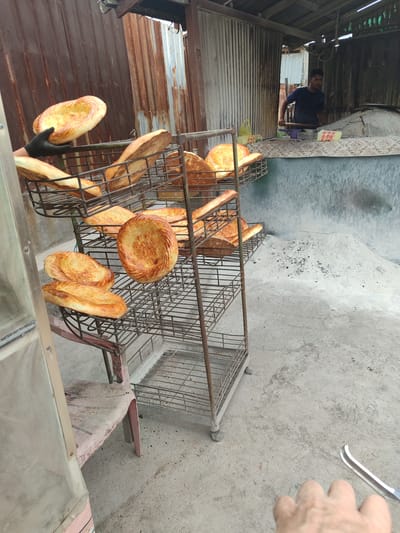 The image depicts an outdoor bakery in Tokmok City, Kyrgyzstan, where flatbreads are being prepared. A man wearing a dark blue t-shirt is visible in the background, actively working near a large, domed oven, likely a tandoor. He is holding a long-handled tool, possibly for placing or removing bread from the oven. In the foreground, a wire rack holds several freshly baked flatbreads, showcasing their golden-brown color and characteristic circular or elongated shapes. One person's gloved hand is visible holding a flatbread, suggesting it is either being presented or moved. The environment appears to be a rustic setting with corrugated metal structures and a concrete floor, typical of many local businesses in the region. The lighting suggests it is daytime.