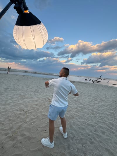 The image depicts a scene at a beach in Novo Oriahovo, Bulgaria. A man wearing a white collared shirt, blue and white striped shorts, and white sneakers is standing on the sand. He appears to be in the middle of a movement or action. The beach is sandy with some driftwood visible. A light attached to a tripod stands to the left. The sky is partially cloudy, with colors suggesting either sunrise or sunset. In the background, another person is walking along the shoreline.