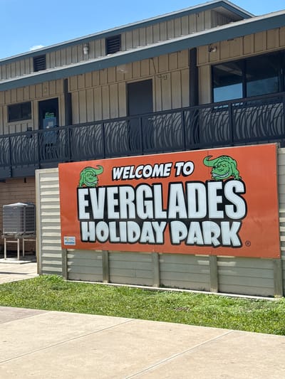 The image shows the entrance to Everglades Holiday Park in Fort Lauderdale, United States. An orange sign displays "WELCOME TO EVERGLADES HOLIDAY PARK" with alligator graphics. The sign is positioned in front of a building with a gray roof and beige siding. Decorative black metal railings are visible. To the right of the sign is a patch of grass and a concrete walkway. An air conditioning unit sits underneath the sign. The sky is visible in the upper left corner.