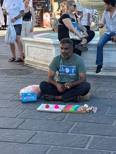 A man sits cross-legged on a cobblestone street, holding a cigarette, in Chania, Greece. He wears a green t-shirt with a blue emblem on it, dark pants, and sandals. In front of him, a small white board displays a red toy tomato and a pink toy heart. To the left of the board, a blue toy box with pictures of animals and a red and white striped bag are placed on the ground. In the background, a stone fountain with water flowing is visible, and several people are seated around it. One woman in a black top and white pants stands near a fountain, holding a shopping bag, and another woman with short blonde hair wears sunglasses and a black dress. The environment appears to be a public square or pedestrian area, typical of a tourist destination.