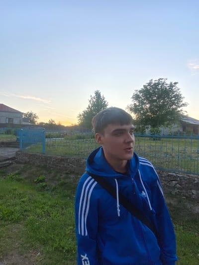 An outdoor, waist-up, medium shot captures a young man standing in front of a blue fence in the village of Tovstoluh, Ukraine, during sunset. The man, with short dark hair and a fair complexion, wears a bright blue hooded tracksuit with white stripes on the sleeves and a black bag slung across his chest. He is looking to his right, with his mouth slightly open as if speaking. The background features a stone wall, a blue metal fence with decorative elements, green grass, trees, and houses with red roofs, characteristic of a rural Ukrainian setting. The sky is a gradient of pale blue to orange due to the setting sun, with wispy clouds. The lighting suggests it is late afternoon or early evening.