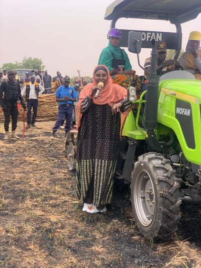 The image shows a woman in a black and white dress and a peach-colored headscarf holding a microphone and speaking in front of a green tractor with the word "WOFAN" written on the side. The tractor is parked in a field that appears to be recently burned. Several men are standing in the background behind the tractor, including a woman wearing a green shirt and a purple cap. The woman is standing in front of the tractor near a field with burned grass in the background. The location is Ringimawa, Nigeria, at coordinates (latitude: 11.666, longitude: 8.416).