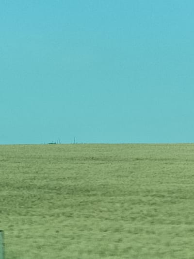 A distant, hazy line of industrial structures and possibly wind turbines is visible on the horizon, appearing as dark silhouettes against a bright, clear, turquoise sky. The foreground is dominated by a vast, flat field of dry, light green grass or crops, showing subtle undulations. The overall impression is one of a very wide-open, rural landscape under a bright, possibly hot, day. The low resolution and distance of the industrial elements suggest they are many kilometers away, possibly near Třemošnice, Czechia, and atmospheric conditions are creating a mirage-like effect. No people are visible, and the scene is devoid of any discernible activity or text.