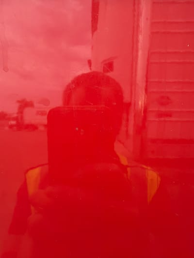 The image is a reflection on a red, glossy surface, possibly a vehicle. A person is visible in the reflection, holding a smartphone and wearing a safety vest. In the background, a building or structure with horizontal lines, possibly a trailer or part of a building, is reflected, along with some indistinct shapes that could be trees or other vehicles. The overall scene suggests an outdoor, possibly urban or industrial, setting in Murfreesboro, United States of America.