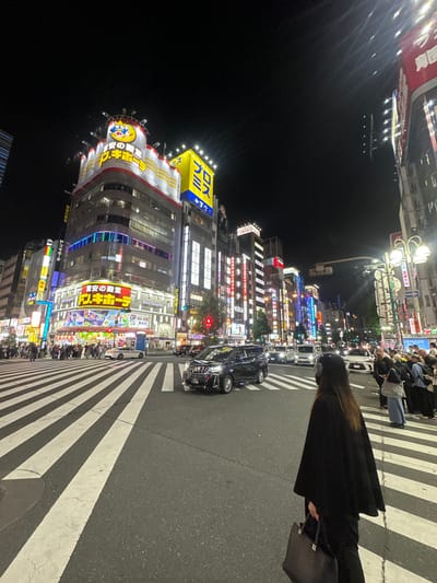 The image shows a street scene at night in Shinjuku, Japan. The foreground features a crosswalk with thick white stripes on an asphalt road. A person wearing a black coat and carrying a black bag is partially visible on the right. Several black taxis are stopped on the road near the crosswalk. A crowd of people can be seen further down the street.

Tall buildings with brightly lit signs and advertisements line the street. A distinctive building with a yellow and blue Don Quijote sign stands out. Other signs and advertisements are in Japanese. The lighting suggests a vibrant and bustling urban environment.