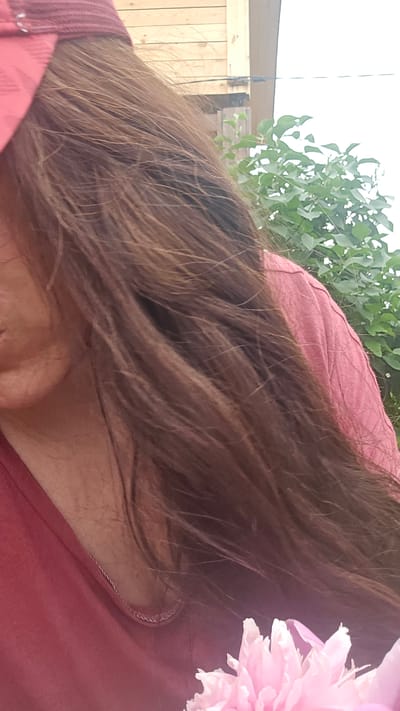 The image captures a close-up view of a person with long, brown hair, wearing a pink t-shirt and a maroon baseball cap. The person's left cheek and part of their chin are visible. The background features wooden siding of a building and lush green foliage, suggesting an outdoor setting. The lighting is bright, indicating daytime. The image appears to be taken in Ермолаевский Затон, Russia, given the context provided. The lower right corner of the image shows a blurry pink flower.