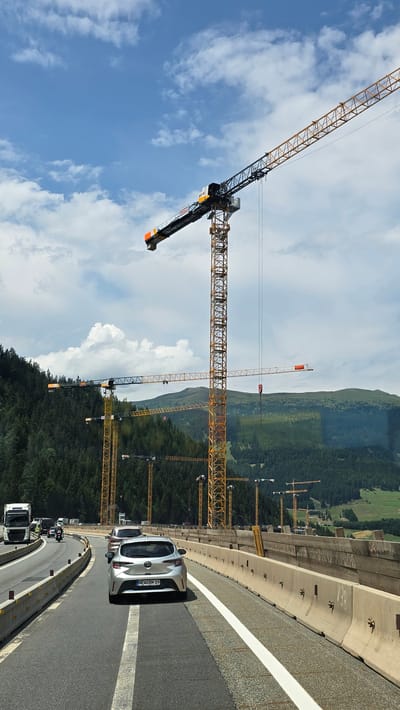 This is a vertical, eye-level, medium shot of a construction site with multiple tower cranes, likely in Gries am Brenner, Austria. The scene is set against a backdrop of lush green mountains and a bright blue sky with scattered white clouds. A paved road curves through the foreground, with a white lane marker and a concrete barrier. Two silver cars are visible on the road, one of which has a license plate that reads "NEW BK 35". Several construction vehicles, including a white truck, are also present on the road further down. The numerous tower cranes dominate the middle ground, suggesting a significant construction project is underway. The time of day appears to be daytime, with clear and sunny weather conditions. The overall impression is one of industrial activity within a scenic natural environment.
