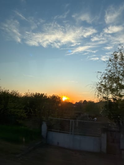 The image captures a vibrant sunset over a rural or semi-rural landscape. The sky dominates the upper portion, transitioning from a clear, light blue on the left to warm hues of orange, yellow, and soft pink towards the right, where the sun is setting. Wispy, white cirrus clouds streak across the blue part of the sky, catching the light.
The sun, a bright and intense golden orb, is positioned on the right side of the horizon, partially obscured by a dense line of silhouetted trees and bushes. Its powerful light creates a distinct sunburst effect, with bright rays extending outwards. There are noticeable lens flares or reflections visible in the sky, particularly a bright blue one in the mid-left area, suggesting the photo might have been taken through a window or glass.
Below the horizon, the landscape is darker, featuring more trees and vegetation. In the bottom right section, faintly visible are what appear to be some man-made structures: possibly a fence or railings, and a larger, darker structure that could be part of a building or wall. A small, light-colored, round object, possibly a football, is discernible in the distant grass area on the right. Blurred tree branches are visible along the top edge of the frame, hinting at foreground elements. The overall impression is one of a peaceful evening scene in Glojene, Bulgaria, during the golden hour of sunset. There is no visible text.