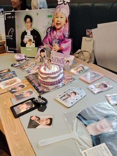The image depicts a table decorated with K-pop merchandise, specifically for an artist named "WONWOO." The setting appears to be a casual gathering or fan event.
Dominating the background is a large cardboard cutout featuring two images of WONWOO. The top image shows him wearing glasses and holding a sign that says "HAPPY BIRTHDAY." Below him is a smaller cutout of a child in traditional Korean attire, smiling. The cutout also has a "REC 06:13" graphic and a date "09/19/2026."
In the foreground, the table is laden with various items:
* A two-tiered cake with purple frosting.
* Numerous photocards and small framed photos of WONWOO.
* Several clear plastic photo holders with pictures of the artist.
* A keychain with a plush toy and a heart-shaped charm.
* A hand fan with a large image of WONWOO.
* A gift bag is partially visible in the background.
The lighting suggests an indoor setting, possibly a cafe or a private room, with the overall atmosphere focused on celebrating WONWOO. The presence of the "09/19/2026" date on the backdrop might indicate a future event or a symbolic date.
There are no explicit details in the image that pinpoint the location to Serang, Indonesia. The provided information about the city of Serang, Indonesia, cannot be confirmed or refuted based on the visual content of the image.