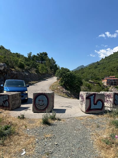 The image shows a blue van parked on a winding road next to a steep, tree-covered hillside. The road is bordered by concrete blocks covered in graffiti, including a smiley face and a stylized letter "F". The hillside is lush with green trees, and a building with a red roof is visible higher up the slope. The sky is clear and blue, with a few white clouds. The sun is shining brightly, casting shadows on the road and the concrete blocks. The overall scene suggests a sunny day in a mountainous or hilly region. There is no visible activity taking place in the image beyond the parked van. The graffiti on the concrete blocks are the only visible text.