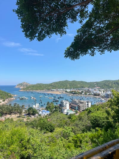 A high-angle, full shot captures a vibrant coastal town nestled in a bay. The sky is a clear, bright blue, with scattered white clouds. In the foreground, lush green trees frame the scene from the top, and a yellow-and-black metal barrier runs along the bottom right.
The bay itself is dotted with a multitude of boats, varying in size and color, anchored in the turquoise water. These boats range from small, traditional fishing vessels to larger, more modern craft. Some appear to be docked near a pier or floating structures.
Along the shoreline of the bay, a town sprawls, characterized by a mix of buildings. There are low-rise structures, some appearing to be residential homes, and several taller buildings, including multi-story hotels or apartment complexes. Some buildings are under construction, with exposed concrete frameworks.
The town is backed by rolling green hills covered in dense vegetation, creating a picturesque backdrop. The coastline curves around the bay, with a rocky outcrop visible on the left side, also covered in greenery. The overall impression is a bustling coastal community during daylight hours, likely a popular tourist destination within Vĩnh Hải, Vietnam.