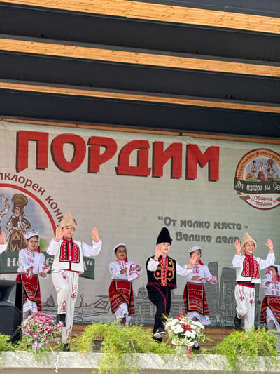 This is a medium shot of a group of children performing traditional Bulgarian folk dances on an outdoor stage. The children are dressed in colorful, embroidered costumes. The stage is decorated with a large banner that reads "ПОPДИM" in red letters, a Cyrillic word for Pordim, a town in northern Bulgaria. The banner also features a circular emblem with images of folk dancers and text in Bulgarian. Some of the children are waving at the audience. In the foreground, there are arrangements of flowers and greenery. The background of the stage depicts a printed scene of buildings and a landscape. The performance is likely part of a cultural festival or event in Pordim, Bulgaria.