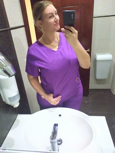 A woman with blonde hair pulled back is taking a mirror selfie in a bathroom. She is wearing a purple scrub top and pants. She has several rings on her fingers and a watch on her left wrist. She is holding a black smartphone in her right hand. In the background, there is a white sink with a chrome faucet, a soap dispenser, a trash can, and a wooden door. The walls are tiled with a pattern of dark and light stripes. The floor is dark tile. The image appears to have been taken in Yagoda, Bulgaria.