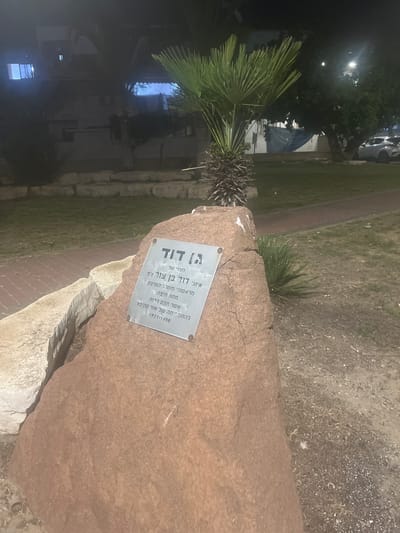 This media file shows a memorial plaque attached to a large rock in a park-like setting at night. The plaque, written in Hebrew, commemorates someone named David Ben Tzur, who was among the founders of the Housing Ministry in the Haifa district and who "built Or Akiva." The dates 1921-1998 are inscribed below his name. A palm tree is visible to the left of the rock, and in the background, buildings and parked cars are dimly lit, suggesting an urban or suburban environment. The lighting indicates it is nighttime, and the overall atmosphere is somber, fitting for a memorial. The stone itself appears to be a rough, natural rock formation, with some lighter-colored stones at its base.