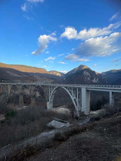 The image displays the Đurđevića Tara Bridge in Đurđevića Tara, Montenegro, a multi-arch concrete bridge spanning a deep, forested river canyon. The light grey bridge features five prominent arches supporting a flat roadway where several vehicles are visible as small light-colored forms. The surrounding landscape consists of rugged mountains under a clear blue sky with scattered white cumulus clouds. Hills on the left exhibit sparse, brown deciduous vegetation under partial sunlight, while mountains to the right and in the background are darker, showing exposed rock faces and patches of evergreen forest. Beneath the bridge, the canyon floor is covered with dormant, dark-branched trees and shrubs. A small, rectangular building with a light-colored roof is situated on the slope below the right section of the bridge, near dark utility lines descending the hillside and a visible dirt path. No individuals are discernible in the scene.