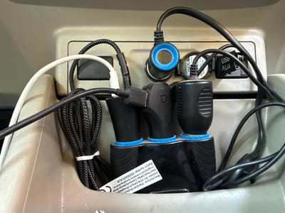The image shows a close-up of a car's center console, filled with various charging adapters and cables. The primary objects are multiple black USB car chargers, some with blue trim, plugged into what appears to be a cigarette lighter socket and a USB/AUX port. A white USB cable and a black braided cable are also visible, connected to the chargers. One of the chargers has a blue ring around its opening. A small black adapter with "USB AUX" written on it is plugged into a port. There's also a circular blue and black adapter with text visible that includes "NOTICE" and "KEEP BATTERY FROM RUN OWN." The background is a neutral-toned plastic interior of a vehicle. The scene is focused on the technical accessories for charging devices within a car. No people are visible, and the lighting suggests an indoor or shaded environment. The overall impression is one of a practical, albeit somewhat cluttered, setup for powering electronics on the go. The visible text on a label identifies the manufacturer as "Shenzhen Shuotuo" and provides an address in China, along with a "Batch number: 20250420JKJA."
