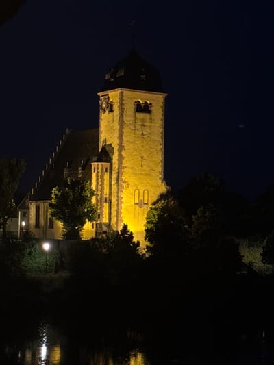 This is a nighttime photograph of a historical church in Hanau, Germany, with the tower illuminated by warm yellow lights. The church has a tall, square tower with a dark, pointed roof and a clock face. Adjacent to the tower is the main body of the church, characterized by a steeply pitched dark roof with several dormer windows. The building is made of light-colored stone, with darker accents and trim. In front of the church, there is dense foliage and trees, some of which are also illuminated by the artificial lights, creating a dramatic silhouette against the dark sky. A single lamppost with a bright light is visible in the foreground, illuminating the bushes. The lower part of the image shows a body of water, likely a river, reflecting the lights of the church and the lamppost. The overall scene conveys a sense of historical significance and peaceful nighttime ambiance.