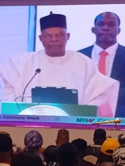 A blurry photograph captures an indoor public speaking event, likely a conference or public address in Tudun Nupawa, Nigeria. The central figure is a man of medium complexion, wearing traditional white attire, including a flowing white robe and a dark, possibly black, cap. He stands behind a podium equipped with a microphone and a tablet device. His expression is serious or focused as he addresses an unseen audience.
Behind and slightly to the right of the main speaker, another man stands, dressed in a dark suit with a white shirt and a red tie. He has a neutral expression. The foreground shows the backs of several audience members' heads, indicating they are seated and facing the speaker. Some audience members wear caps, including one bright yellow cap.
Visible text on the green banner or screen at the podium includes "...PRESIDE..." (likely "PRESIDENT" or "PRESIDENCY") and "...Relations Week." Below this, on a purple-blue strip, the acronym "NRW" is clearly visible, followed by what appears to be part of another word starting with "C" (e.g., "Conference"). A golden, possibly ceremonial, staff or emblem is also partially visible on this lower strip. The overall scene suggests a formal event, possibly governmental or an important organizational meeting, given the prominence of the speaker and the "PRESIDE" text.
