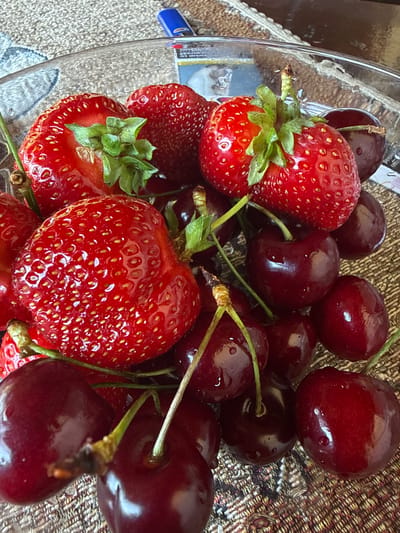 A close-up, top-down shot captures a clear glass bowl filled with fresh strawberries and dark red cherries, resting on a textured, woven placemat. The fruits are glistening, suggesting they are recently washed. Some strawberries have green leafy tops. The lighting is bright, highlighting the vibrant red of the fruits and the reflective surfaces of the bowl and the water droplets on the cherries. A blue lighter is visible in the upper background. The scene is likely in Rudartsi, Bulgaria, given the context of the location.