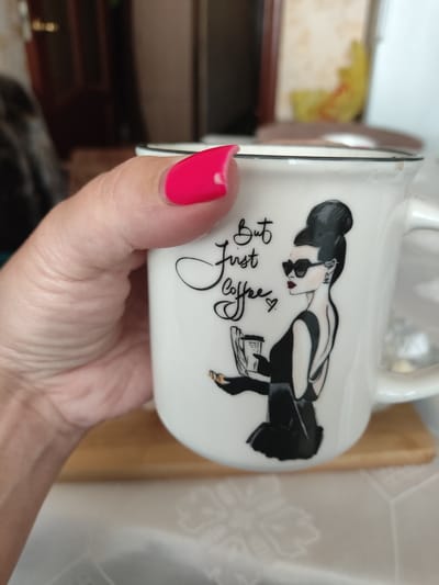 In this close-up, indoor shot, a right hand with a bright pink fingernail is holding a white ceramic mug. The mug features a black and white illustration of a stylized woman in a black dress, sunglasses, and a high bun, holding a coffee cup and what appears to be a newspaper. Text on the mug reads "But First Coffee". The background is blurred, showing hints of furniture and possibly a window, suggesting a domestic setting. There is no indication of Myadzyel, Belarus, in this image.