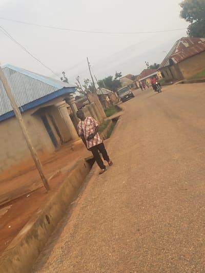 This is a street-level photograph taken in the city of Gumel, Nigeria. The scene depicts a street with buildings on either side and a few people and vehicles present. In the foreground, on the left, is a man wearing a checkered shirt and dark pants. He is standing on the edge of a drainage channel. To the right of the street, there are several buildings with corrugated metal roofs, typical of residential structures in the region. In the background, several other people can be seen walking along the street, and a motorcycle is visible. The overall environment appears to be a typical rural or semi-urban setting in Nigeria, with a dusty road and modest buildings. The sky is overcast, suggesting a cloudy day. The photograph has been taken at an angle, giving it a dynamic perspective.