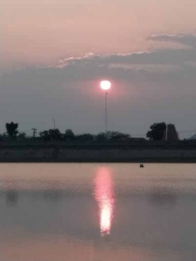 The image depicts a sunset over a body of water, likely a reservoir or a lake, in Sambirejo, Indonesia. The sun, a bright pinkish-red orb, is partially obscured by thin clouds in the upper center of the frame. A reflection of the sun streaks down the water's surface, creating a shimmering path of light. Along the far bank, silhouetted against the dusky sky, are trees and structures. One prominent structure is a tall, slender tower topped by a small circular object, likely a communication tower. To the right, a building with a distinctive pyramid-shaped roof, possibly a temple or a unique architectural feature, is visible. In the middle distance on the water, a small dark object, appearing to be a boat with two occupants, is present. The overall atmosphere is tranquil and serene, characteristic of a sunset.