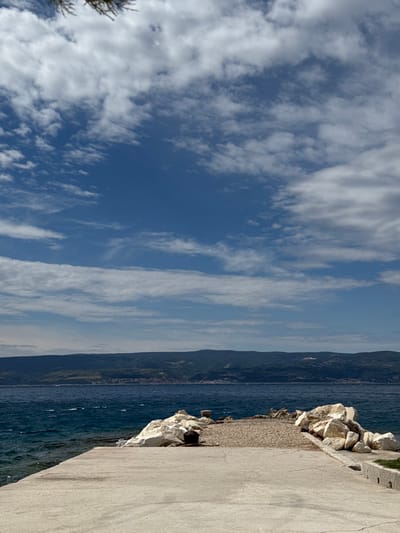 The image shows a concrete pier in the town of Dugi Rat, Croatia, located at the coordinates 43.446 latitude and 16.635 longitude. The pier is empty with white rocks on either side. The water is blue and choppy. There are large white clouds in a bright blue sky. Behind the water is a distant mountain range with a small town in the foreground of the mountains. The scene is wide and expansive. There are no people visible in the image. The sun is shining and the weather is pleasant. It is a daytime scene.
