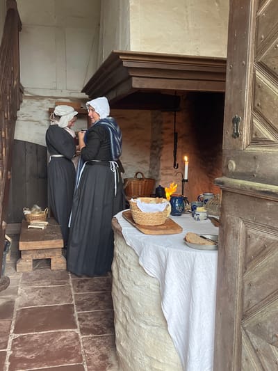An interior scene in Staudernheim, Germany, depicts two adult females in period attire interacting within a rustic room. The woman on the right wears a dark gown, a white apron, a white bonnet, and a blue and white striped shawl, facing left and slightly towards the second individual. The second woman, positioned to the left, wears a dark gown, a white apron, and a light-colored bonnet, facing right towards the first. Both individuals are standing and appear engaged in a conversation.

The foreground right displays a partially open, ornate wooden door featuring a dark metal latch and paneled design. Centrally, a round, rough-hewn stone table is draped with a white linen tablecloth. Resting on the table are a wicker basket containing bread, a wooden cutting board, a metal dish holding a slice of bread, a blue ceramic jug, a small white and blue ceramic pot with butter, two ceramic mugs, and a clear glass pitcher. Behind the table, a lit white taper candle in a black metal holder illuminates the area. Further back, a large, deep fireplace contains a small, active fire, emitting an orange glow; a dark metal hook hangs within the hearth. A robust wooden mantelpiece is positioned above the fireplace.

To the mid-left, a rustic wooden bench supports a small wicker basket containing yarn or fibers, alongside several bundles of dried reeds or straw. In the background left, a wooden staircase with an open railing ascends. The room's architecture includes light-colored, rough stone walls, a floor paved with dark reddish-brown square tiles, and exposed wooden structural elements. Lighting is provided by ambient light from an unseen source and the warm illumination from the candle and fireplace.