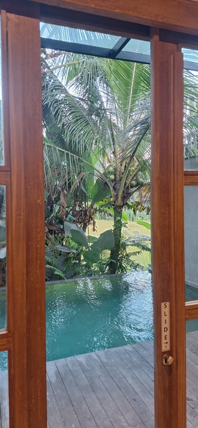 The image shows a view through a wooden doorway onto a tropical scene. In the foreground, there is a wooden deck and a turquoise swimming pool. Beyond the pool, lush green vegetation, including palm trees and ferns, is visible. In the background, a rice paddy field can be seen. On the right side of the doorway, a small sign reads "SLIDE" with an arrow pointing to the right, suggesting a sliding door mechanism. The scene appears to be from a resort or private villa in Payangan, Indonesia, a region known for its natural beauty and rice terraces.