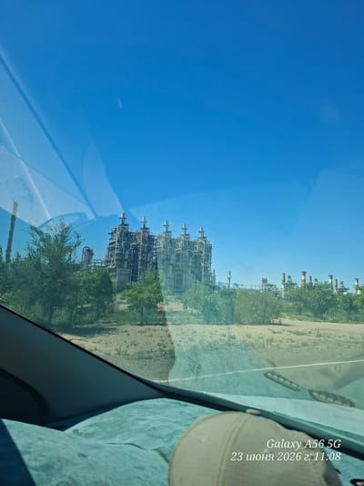 This is a photograph taken from inside a vehicle, looking through the windshield at an industrial complex under a clear blue sky. The complex appears to be a refinery or chemical plant, with numerous towers, pipes, and stacks. Some of the stacks are emitting smoke or steam. Green trees and vegetation are visible in the foreground and around the industrial structures. The lower part of the image shows the dashboard and a tan baseball cap on a light blue surface, likely a seat cover. A timestamp in the lower right corner reads "Galaxy A56 5G 23 июня 2026 г. 11:08". The location is identified as Kyrkkyz, Uzbekistan.