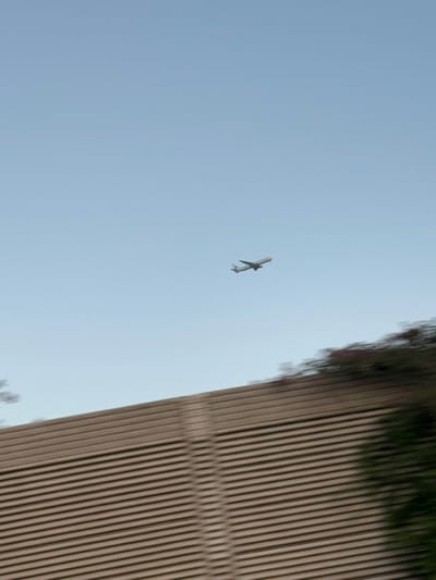 The image displays a light-colored commercial airplane in flight against a clear, light blue sky over Rosh HaAyin, Israel. The aircraft is positioned in the upper-right quadrant, appearing to be in an ascent phase with its landing gear extended. The lower third of the frame is occupied by a large, horizontally ribbed, light-brown or beige barrier structure, which exhibits significant motion blur, suggesting the photograph was taken from a moving vehicle. Above and to the right of this structure, blurred green and red foliage is visible, also affected by motion blur. No individuals are depicted.