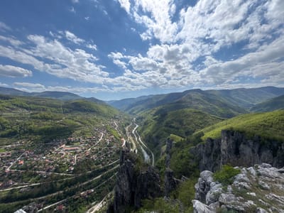 The image captures a broad, scenic view overlooking Milanovo, Bulgaria, from a high vantage point. In the foreground, rocky terrain and jagged cliffs are visible, suggesting a mountainous or elevated location. A small town sits nestled among green hills and forests to the left, with houses and buildings clustered together and traversed by winding roads. A river or stream meanders through a valley, flanked by roadways or railway lines that run alongside it. The background features rolling hills and mountains fading into the distance. The sky is blue and filled with fluffy white clouds, suggesting a clear, sunny day. The scene depicts a natural landscape combined with human habitation and infrastructure.
