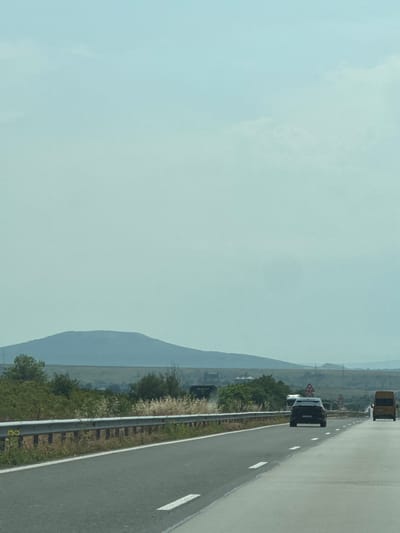 This is a landscape image taken from a moving vehicle on a highway, likely in the vicinity of Novi Pazar, Bulgaria. The scene is dominated by a vast, pale blue sky with wispy clouds. In the middle ground, a line of low, rolling hills and vegetation stretches across the frame. A prominent, rounded mountain rises in the distance, its contours softened by the haze. The foreground features a two-lane asphalt highway with white dashed lane markings. A metal guardrail runs along the left side of the road, separating it from the grassy embankment and scattered green bushes. Several vehicles are visible on the highway: a black SUV in the middle lane, a yellow van on the right lane, and other vehicles further ahead, appearing as blurs. Traffic signs are present, including a speed limit sign showing "90" and another showing "70". The overall impression is one of a journey through a rural, open landscape under a hazy sky.