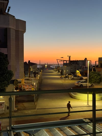 This image captures a waterfront scene in Gafanha da Nazaré, Portugal, during twilight or early evening. The sky exhibits a clear gradient, transitioning from a vibrant orange and yellow hue at the horizon to a pale blue overhead, with a single faint white point visible in the upper blue.
In the foreground, a dark metal railing with horizontal bars diagonally crosses the bottom portion of the frame. Below this railing, a partially visible, ribbed, light-colored structure, possibly an awning or sunshade, is present.
The midground features a wide, paved promenade or street extending into the distance, illuminated by multiple warm-toned streetlights that cast long shadows. On the left, a multi-story concrete building with vertical ribbed detailing is prominent. Below it, a dense green bush partially obscures a circular, illuminated sign displaying "Nova Barr's." An outdoor dining area with numerous small, square tables and orange chairs is arranged beneath yellow awnings, lit by artificial light.
On the right side of the promenade, a white sedan and a silver hatchback are parked. Adjacent to the white sedan, a small, round, leafy green tree stands beside a streetlight. A low, curved, light-colored barrier borders the promenade on the far right.
One individual, dressed in a dark jacket and lighter trousers, walks away from the viewer on the right side of the promenade, casting an elongated shadow to their left. Two additional figures are discernible standing near the parked silver hatchback further down the street.
The background shows the promenade leading to a dark body of water, likely the sea or an estuary, with subtle wave patterns. Buildings with angular roofs and various structures, including what appears to be a windsock and a horn-like device on a pole, line the waterfront. Distant lights are visible along structures extending into the water, suggesting jetties or breakwaters.