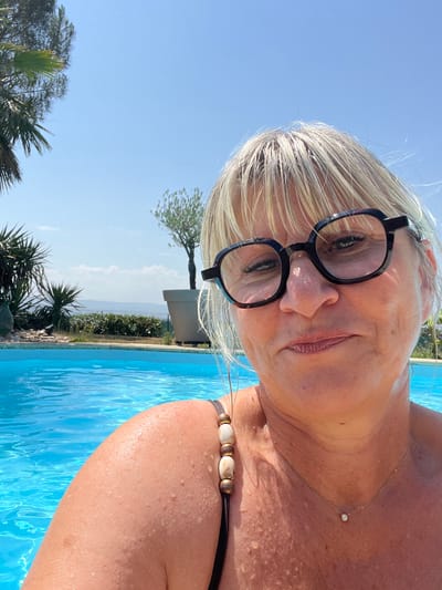 A blonde woman with glasses is in a swimming pool, taking a selfie. She is wearing a swimsuit with decorative beads on the strap. The background shows a clear blue sky, palm trees, and other vegetation. The setting appears to be a private residence or resort. The image was captured in the city of Saint-Galmier, France.