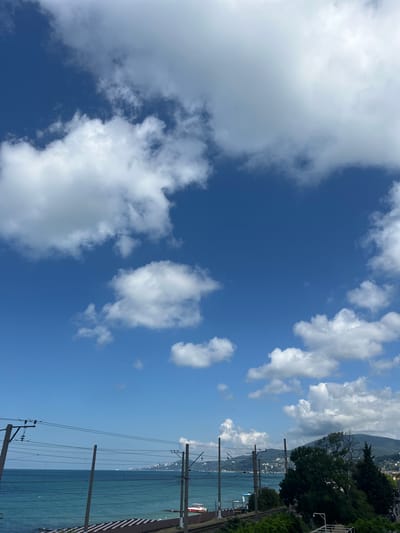 The image captures a view of the Black Sea coast in Khostinskiy, Russia, under a bright blue sky dotted with fluffy white clouds. In the foreground, a set of overhead power lines for a railway track stretches across the frame, supported by concrete poles. Beyond the tracks, the turquoise waters of the sea are visible, meeting a distant coastline with buildings and greenery. A strip of beach with a patterned surface is situated between the railway and the sea. To the right, a lush green hillside rises, populated by trees and vegetation, with some buildings visible on its slopes. The overall impression is one of a sunny coastal day.