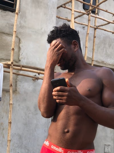 An adult male is depicted outdoors in Enugwu-Ukwu, Nigeria, under bright daylight. The subject possesses dark skin, short, dark, curly hair, and a beard. He is shirtless, revealing a muscular physique, and wears red "O'NEILLS" brand underwear. His right hand covers the upper part of his face, while his left hand holds a black smartphone with a textured case, positioned in front of his torso, indicating interaction with the device. The background consists of a light grey, unfinished concrete or plaster wall, against which bamboo scaffolding is erected. A dark window frame is visible in the upper background, and a white garment hangs on the scaffolding to the far left.