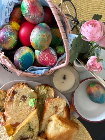 The image depicts an Easter celebration scene. A basket filled with dyed Easter eggs, some with marbled patterns, is visible on the left, along with a small yellow chick. Pink rose flowers in a pot sit adjacent to the basket. Nearby is a lit candle in a glass container. To the right, there is a plate of sliced bread with raisins, some pieces decorated with green icing. An Easter egg in a dish is present. Eyeglasses and a golden woven mat are also present in the background. There is no indication of the location, Bretton, United Kingdom in this picture.