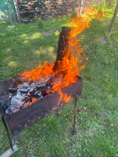 In this media file, a fire burns within a metal grill, which is situated on a grassy area with scattered small white flowers. The grill is long and rectangular with a perforated side. To the left of the grill, a pile of firewood is visible, along with a portion of a green object that could be a tarp or a bin. The fire consists of bright orange flames consuming crumpled paper or cardboard. The scene appears to be outdoors, likely in a yard or park, given the presence of grass and firewood. The lighting suggests it is daytime, possibly late afternoon due to the warm hues of the fire and shadows. There are no people visible in the frame, and no text is present. The overall impression is a rustic, outdoor setting where a fire is being managed, possibly for cooking or for disposal of waste materials. The location context is Zviahel, Ukraine.